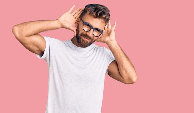 Jonge spaanse man met vrijetijdskleding en een bril die beide handen op het oor probeert te horen gebaar nieuwsgierig naar roddels gehoorprobleem doof