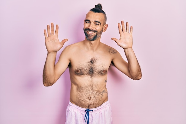 Gratis foto jonge spaanse man shirtless dragen zwempak tonen en omhoog wijzend met vingers nummer tien terwijl hij zelfverzekerd en gelukkig glimlacht.