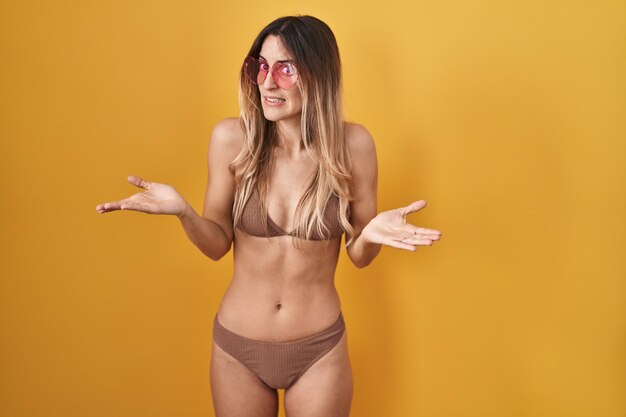 Jonge spaanse vrouw die bikini over gele achtergrond draagt, geen idee en verward met open armen, geen idee concept.