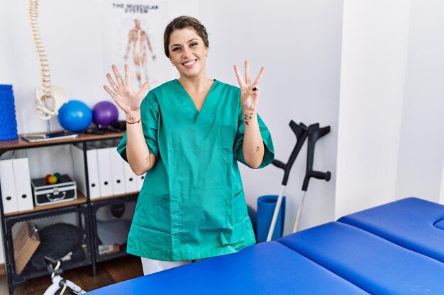 Jonge spaanse vrouw die een fysiotherapeut-uniform draagt en in de kliniek staat en omhoog wijst met vingers nummer acht terwijl ze zelfverzekerd en gelukkig glimlacht.