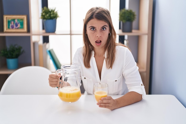 Jonge spaanse vrouw die glas jus d'orange drinkt met een geschokt gezicht en er sceptisch en sarcastisch uitziet, verrast met open mond