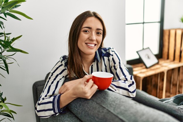 Jonge Spaanse vrouw die koffie drinkt terwijl ze thuis op de bank zit