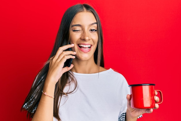 Gratis foto jonge spaanse vrouw die op de smartphone praat en een kopje koffie drinkt knipogend kijkend naar de camera met sexy uitdrukking vrolijk en blij gezicht