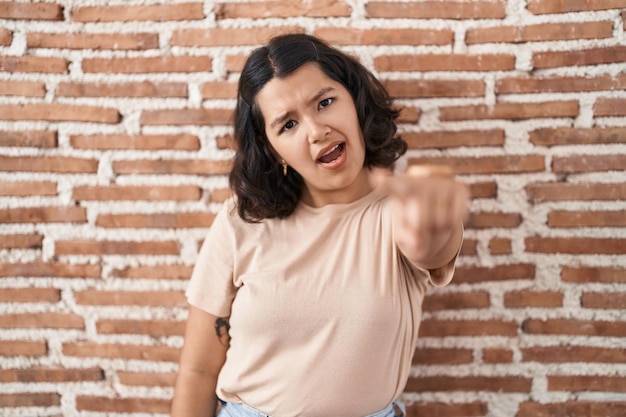 Jonge spaanse vrouw die over bakstenen muur staat en ontevreden en gefrustreerd naar de camera wijst, boos en woedend op jou