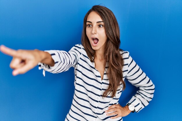 Jonge spaanse vrouw die over een blauwe geïsoleerde achtergrond staat en met de vinger wijst, verrast vooruit, open mond verbaasde uitdrukking, iets aan de voorkant