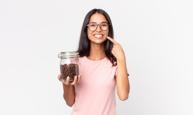Jonge spaanse vrouw die zelfverzekerd glimlacht, wijst naar haar eigen brede glimlach en houdt een fles koffiebonen vast