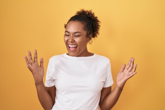 Jonge spaanse vrouw met krullend haar die over gele achtergrond staat en gek en gek viert voor succes met opgeheven armen en gesloten ogen opgewonden schreeuwend. winnaar concept