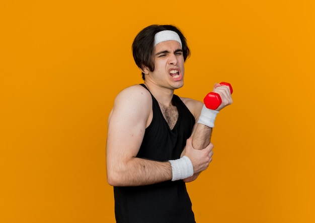 Jonge sportieve man die sportkleding en hoofdband draagt die uitwerkt met halter die verward en ontevreden over oranje muur kijkt