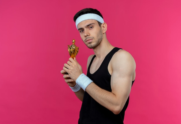 Jonge sportieve man in de trofee van de hoofdbandholding met zelfverzekerde ernstige uitdrukking die zich over roze muur bevindt
