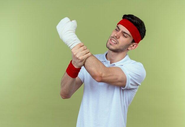 Jonge sportieve man in hoofdband die zijn verbonden hand raakt die pijn voelt die zich over groene achtergrond bevindt