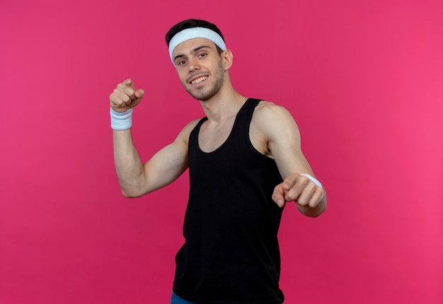 Jonge sportieve man in hoofdband glimlachend wijzend met wijsvingers naar camera over roze