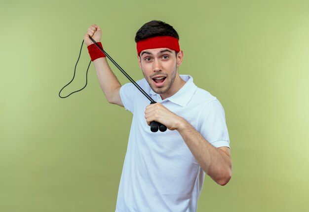 Jonge sportieve man in hoofdband met springtouw kijken camera glimlachend vrolijk permanent over groene achtergrond