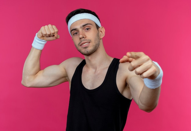 Jonge sportieve man in hoofdband met zelfverzekerde uitdrukking vuist verhogen biceps tonen met wijsvinger naar camera over roze