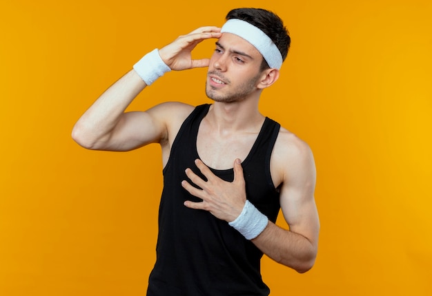 Jonge sportieve man in hoofdband op zoek verward met hand op zijn hoofd voor fout boven oranje