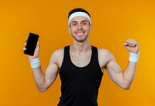 Jonge sportieve man in hoofdband weergegeven: smartphone balde vuist blij en opgewonden staande over oranje muur