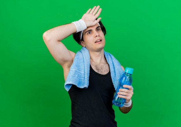 Jonge sportieve man met sportkleding en hoofdband met een handdoek om de nek met fles water op zoek moe en uitgeput