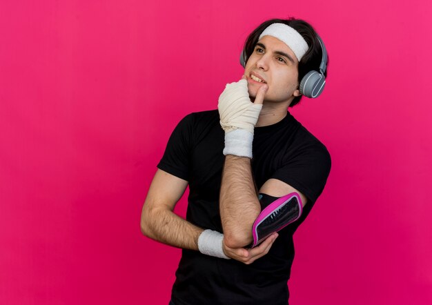 Jonge sportieve man met sportkleding en hoofdband met koptelefoon en smartphone armband opzij kijken met hand op kin denken