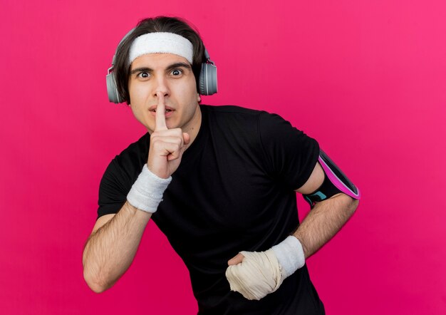 Jonge sportieve man met sportkleding en hoofdband met koptelefoon en smartphone armband stilte gebaar maken met vinger op lippen