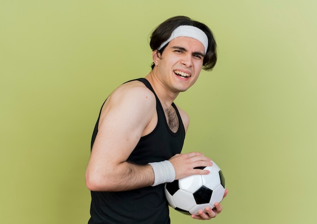 Jonge sportieve man met sportkleding en hoofdband met voetbal lachend verward
