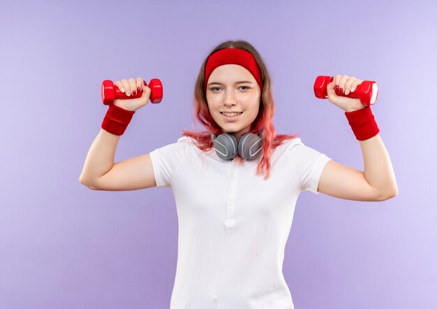 Jonge sportieve vrouw die twee domoren houdt die oefeningen met glimlach op gezicht doen die zich over purpere muur bevinden