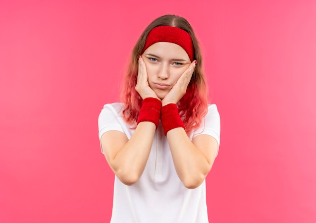 Jonge sportieve vrouw in hoofdband die haar gezicht met wapens aanraakt die moe en verveeld over roze muur kijkt