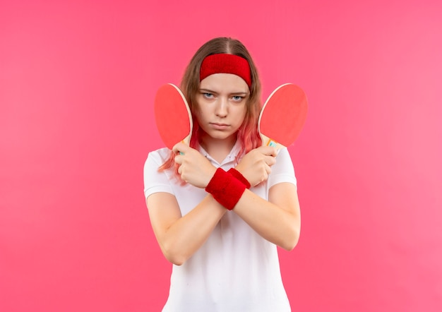 Jonge sportieve vrouw in hoofdband die twee rackets voor tafeltennis houdt die handen kruist met ernstig gezicht dat zich over roze muur bevindt