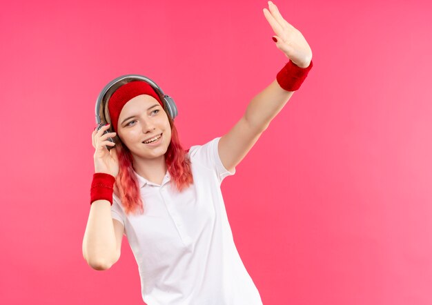 Jonge sportieve vrouw in hoofdband met hoofdtelefoons die opzij gelukkig en positief kijken zwaaien met hand die zich over roze muur bevindt