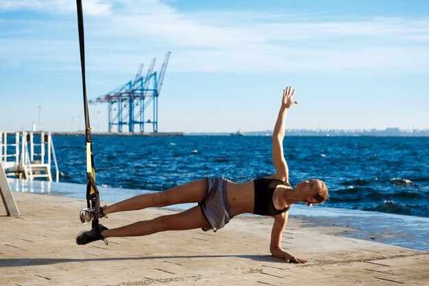 Jonge sportieve vrouw training met trx in de buurt van de zee in de ochtend