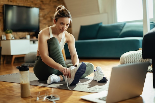 Jonge sportvrouw die laptop gebruikt terwijl ze haar sneakers aantrekt in de woonkamer