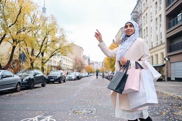 Jonge stijlvolle Arabische vrouw in hijab met boodschappentassen die taxi op straat in de stad probeert te stoppen