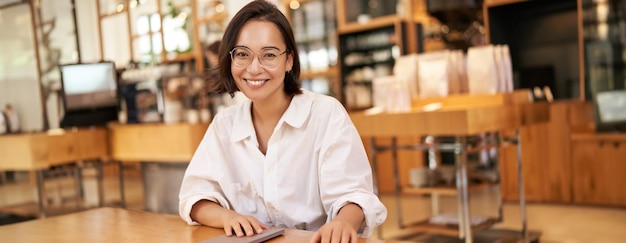 Jonge stijlvolle aziatische vrouw bedrijfseigenaar in glazen zitten in café met laptop glimlachend in de camera