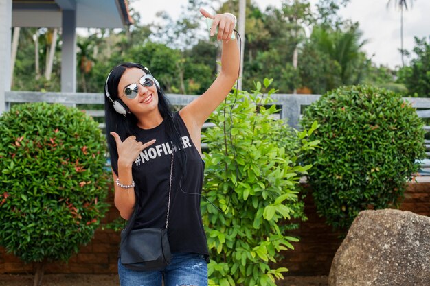 Jonge stijlvolle hipster vrouw in zwart t-shirt, spijkerbroek, luisteren naar muziek op koptelefoon, plezier, poseren, selfie foto nemen op telefoon, vredesteken, grappig gezicht expressie tonen