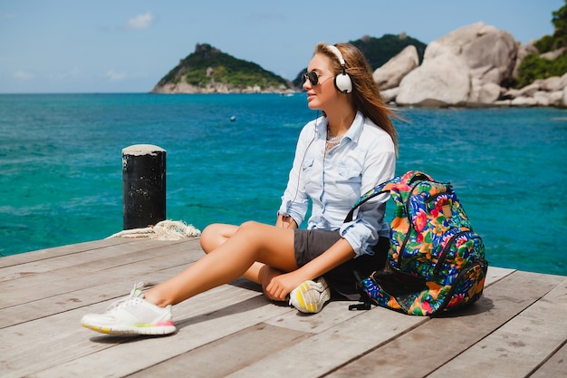 Jonge stijlvolle hipster vrouw reist de wereld rond, zittend op de pier, vlieger zonnebril, koptelefoon, luisteren naar muziek, vakantie, rugzak, denim hemd, gelukkig, tropische eilandlagune