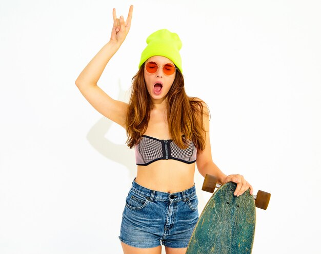 Jonge stijlvolle lachende vrouw model in casual zomer badmode kleding en gele muts poseren met longboard bureau, rock and roll teken tonen. Geïsoleerd op wit