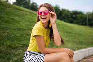 Gratis foto jonge stijlvolle lachende vrouw plezier in stadspark, vrolijke stemming glimlachen, gele top, gestreepte minirok, roze zonnebril, zomer stijl modetrend dragen