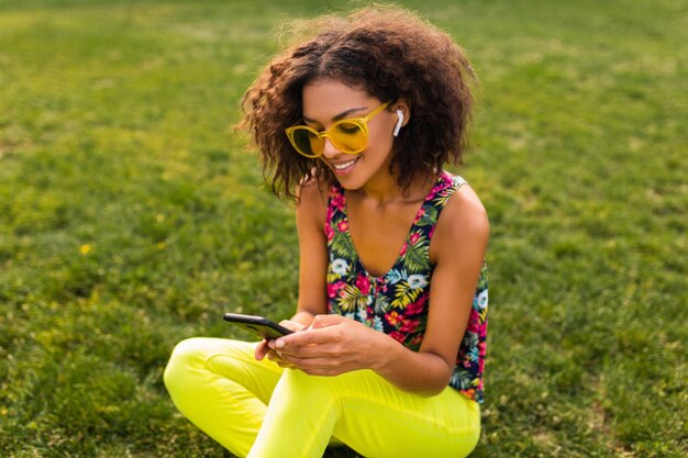 Jonge stijlvolle lachende zwarte vrouw met behulp van smartphone luisteren naar muziek op draadloze oortelefoons met plezier in park