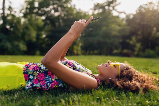 Jonge stijlvolle lachende zwarte vrouw met smartphone luisteren naar muziek op draadloze oortelefoons met plezier in park, zomer mode kleurrijke stijl, liggend op gras, gele zonnebril