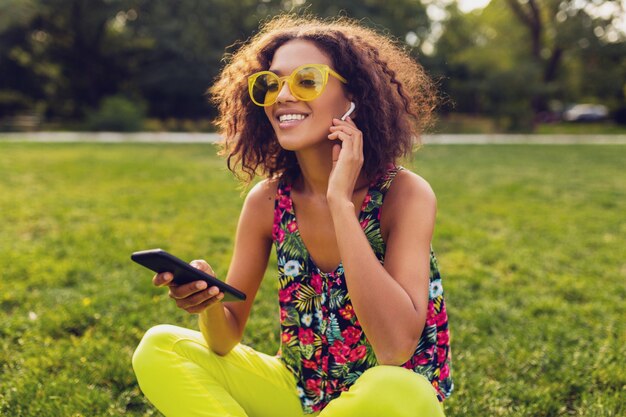 Jonge stijlvolle lachende zwarte vrouw met smartphone luisteren naar muziek op draadloze oortelefoons met plezier in park, zomer mode kleurrijke stijl, zittend op gras, gele zonnebril