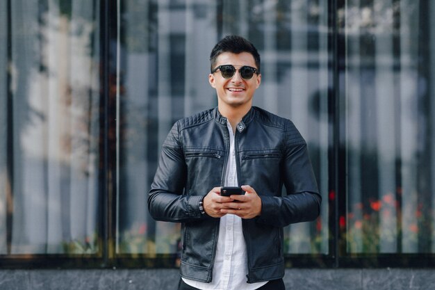 Jonge stijlvolle man in glazen in zwart lederen jas met telefoon op glazen oppervlak