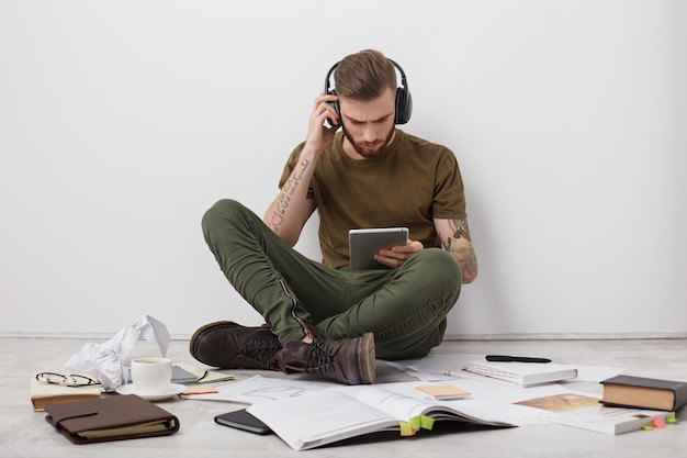 Gratis foto jonge stijlvolle man luistert naar muziek met een koptelefoon, houdt moderne tabletcomputer vast, communiceert online met vrienden of familieleden