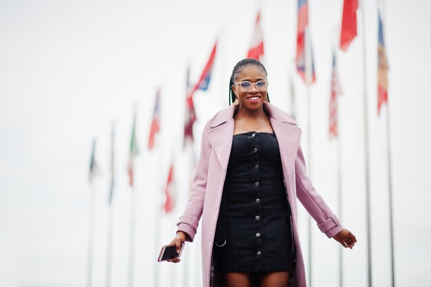 Jonge, stijlvolle, mooie afro-amerikaanse vrouw in straat met een mode-outfitjas tegen vlaggen van verschillende landen van de wereld met een mobiele telefoon bij de hand