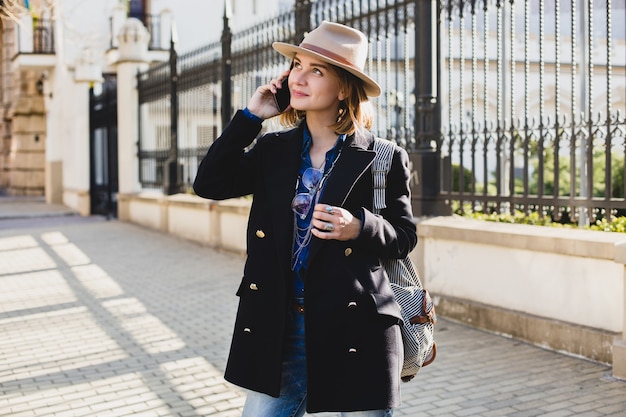 Jonge stijlvolle mooie vrouw glimlachend en praten over haar telefoon, gekleed in donkerblauwe jas en spijkerbroek