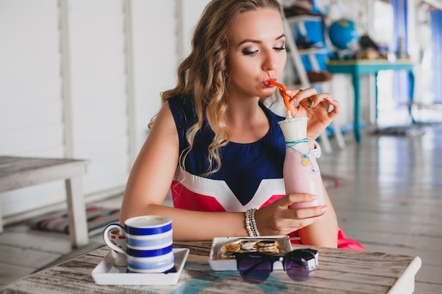 Jonge stijlvolle mooie vrouw in zee café, cocktail smoothie drinken, zonnebril, flirterige, resortstijl, modieuze outfit, glimlachen, mariene kleuren jurk