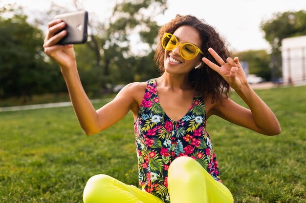Jonge stijlvolle positieve zwarte vrouw selfie foto luisteren naar muziek op draadloze oortelefoons met plezier in park, zomer fashion stijl, kleurrijke hipster outfit