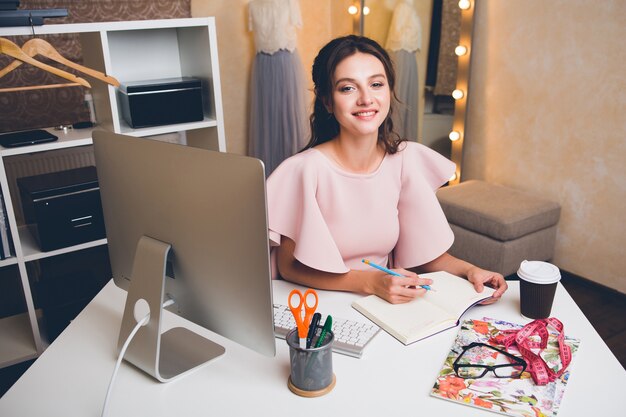 Jonge stijlvolle sexy vrouw in roze luxe jurk zomer trend, chique stijl, modeontwerper werken op kantoor op computer