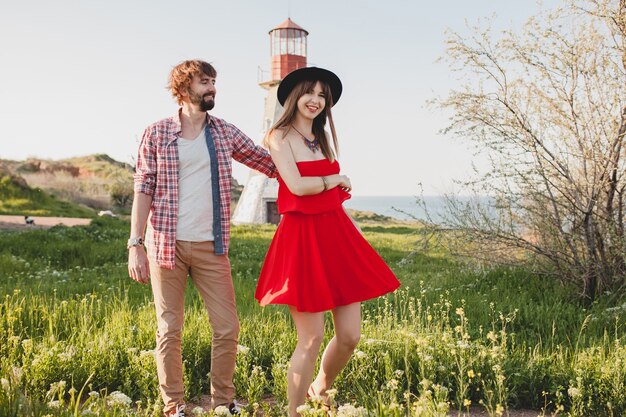 Jonge stijlvolle verliefde paar dansen op platteland, indie hipster bohemien stijl, weekendvakantie, zomer outfit, rode jurk, groen gras, hand in hand