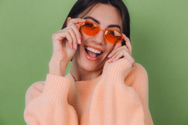 Jonge stijlvolle vrouw in casual perzik trui en oranje bril geïsoleerd op groene olijfmuur gelukkig positief lachend lach rond kopie ruimte