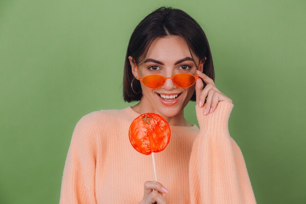 Jonge stijlvolle vrouw in casual perzik trui en oranje bril geïsoleerd op groene olijfmuur met oranje lolly positieve glimlach kopie ruimte