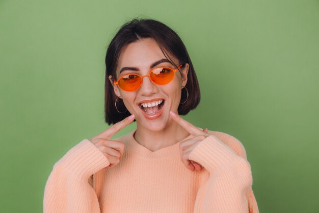 Jonge stijlvolle vrouw in casual perzik trui en oranje bril geïsoleerd op groene olijfmuur positief lachend wijzend op witte tanden met wijsvinger kopie ruimte