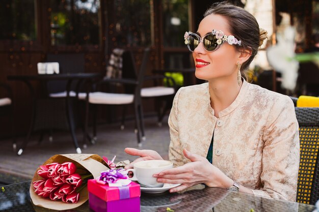 Jonge stijlvolle vrouw, mode zonnebril, zittend in café, kopje cappuccino te houden